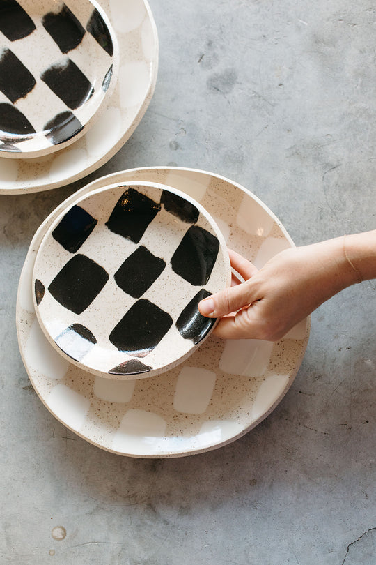 The Object Enthusiast handmade ceramic dinnerware. A hand stacks a black checkered salad plate on top of a larger, white checkered dinner plate.