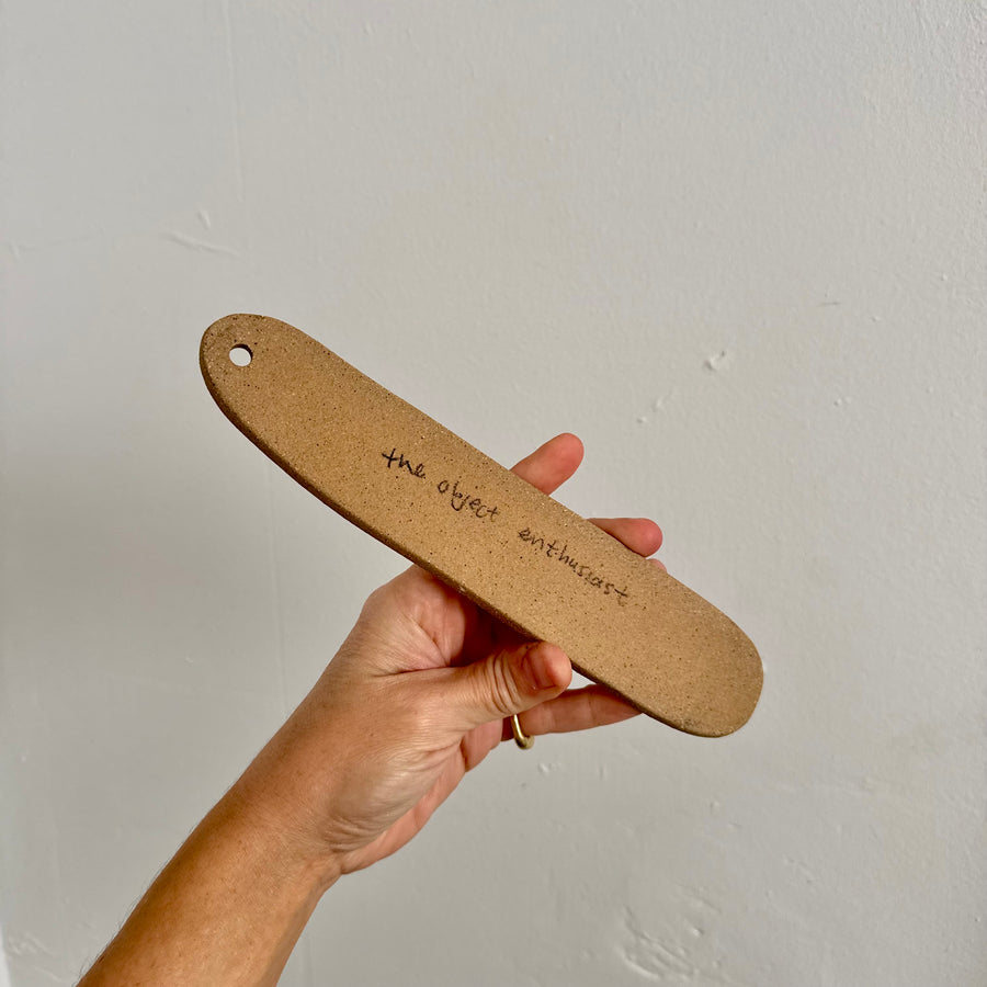 Hand holding a brown ceramic piece with 'the object enthusiast' branding against a plain background
