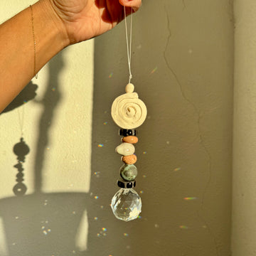 Hand holding a decorative hanging ornament with beads and a crystal prism against a neutral background
