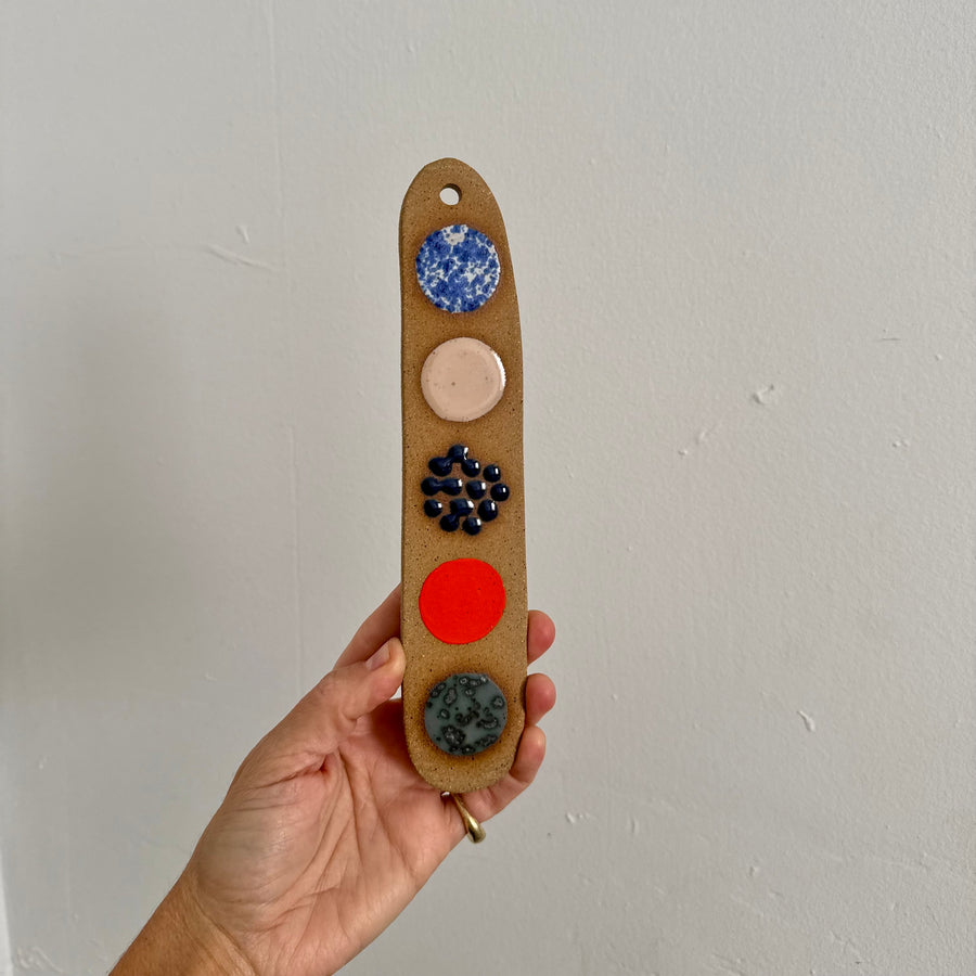 Hand holding a narrow ceramic wall hanging with colorful circles against a plain background