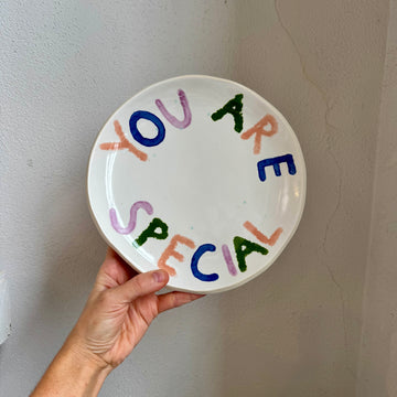 A hand holds a ceramic plate against a white wall. The plate is glazed white and says “you are special” in peach, blue, purple, and green glazes. 