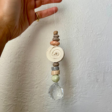 Hand holding a hanging decorative suncatcher with stones and a crystal against a white wall