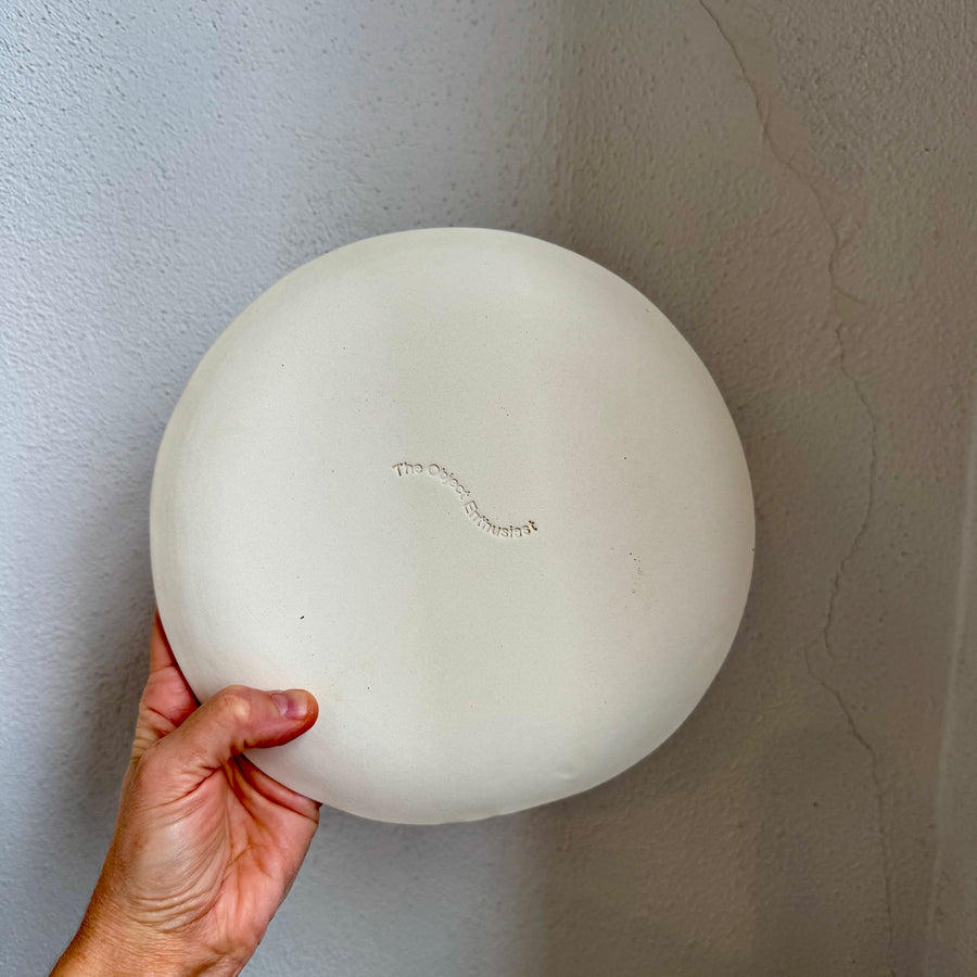 Hand holding a ceramic plate against a plain white wall, this image is the back of the plate showing the logo and the porcelain clay