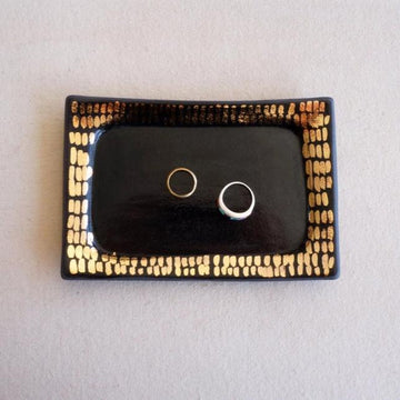 Black and gold rectangular ceramic tray that holds rings and other small pieces of jewelry