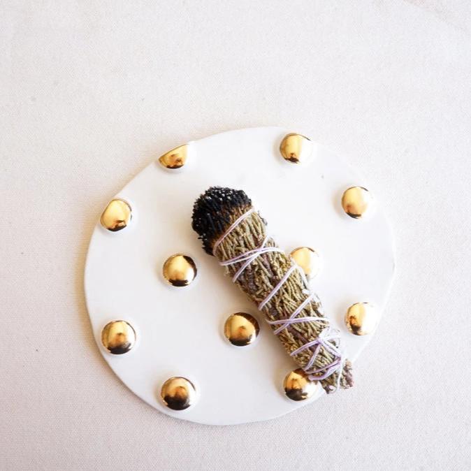 A flat, white and gold polka dotted incense trivet sits on a neutral background with a burned bundle of incense sitting on the trivet