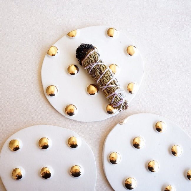 Three flat incense trivets with raised gold polka dots sit on a neutral backdrop one has a bundle of burned incense on it 