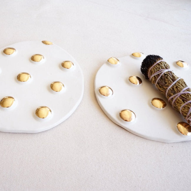 Three white and gold incense trivets in a line, each one is slightly different from the next, one has a bundle of burned incense sitting on it